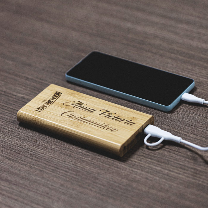 En powerbank i trä med ett namn graverat. En mobil ligger bredvid och laddas.